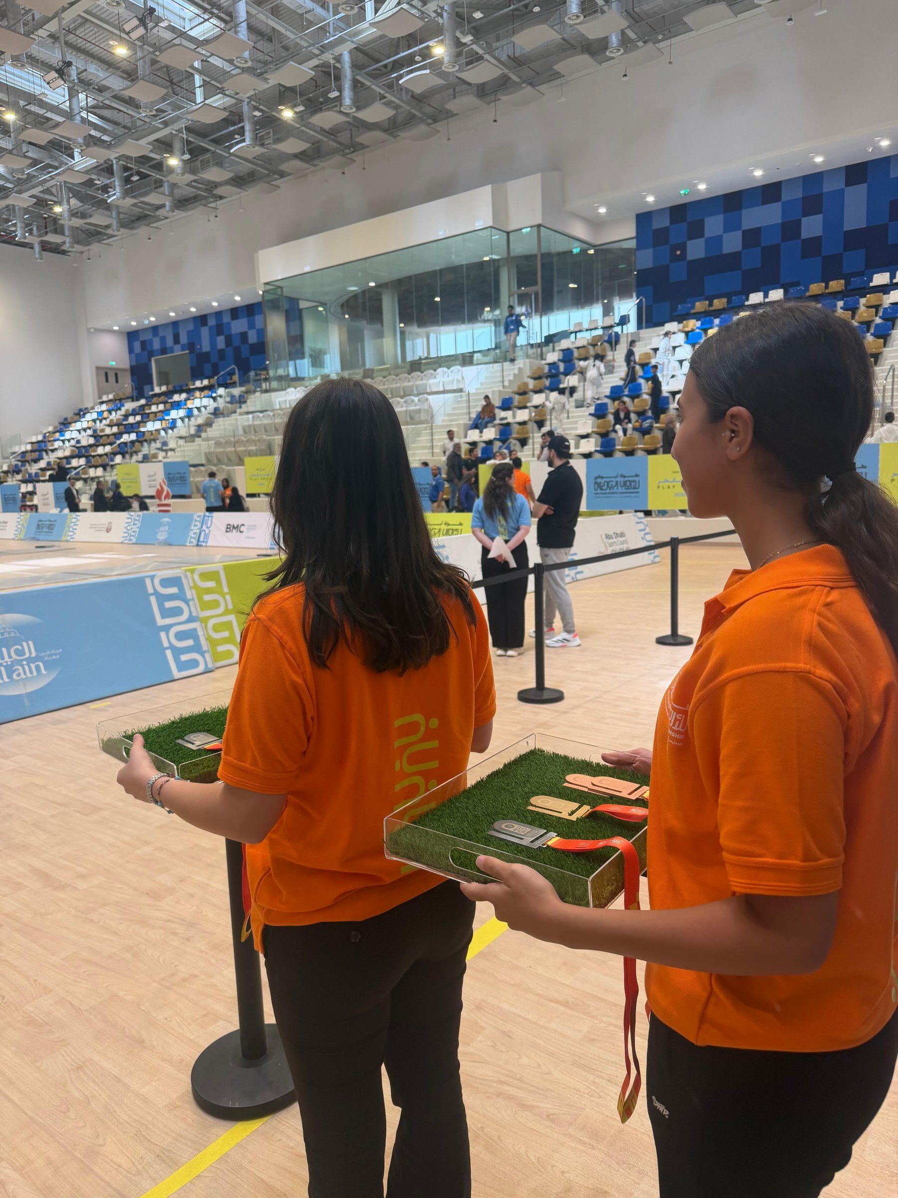 Two hostesses in orange carrying medal trays across the arena
